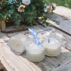 May include: Three clear glass candle jars with lids, two with white candles and one with a blue ribbon and a tag that reads "Olivia's Baby Shower".