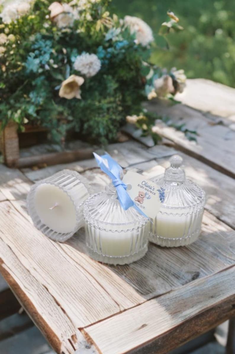 May include: Three clear glass candle jars with lids, two with white candles and one with a blue ribbon and a tag that reads "Olivia's Baby Shower".