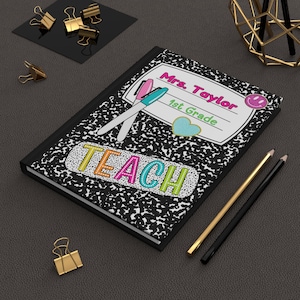 Pode incluir: Um caderno de composição com uma capa salpicada em preto e branco. O caderno tem o texto "Mrs. Taylor 1st Grade" e a palavra "TEACH" em letras coloridas. Dois lápis e clipes dourados também são visíveis.