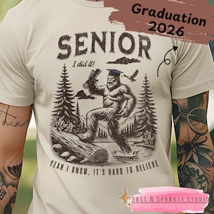 May include: Beige t-shirt with a Bigfoot graphic wearing a graduation cap and holding a diploma. The shirt reads "SENIOR" and "Yeah I know, it's hard to believe." The text "Graduation 2026" is also visible.
