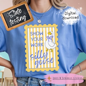 Puede incluir: Camiseta azul claro con un diseño caprichoso de un ganso blanco con un lazo morado, dentro de un marco a rayas amarillas y blancas. El texto dice "Show Your Work Silly Goose". Una pequeña pizarra dice "State testing".