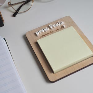 Puede incluir: Un portanotas de madera con una pila de notas adhesivas amarillas. El soporte tiene el texto "Profe Paula enseñando con el" en escritura blanca. Gafas con montura dorada descansan sobre la superficie blanca.