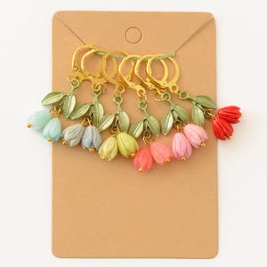 Puede incluir: Una colección de pendientes de flores coloridas exhibidos en una tarjeta marrón. Cada pendiente presenta un aro dorado, hojas verdes y un grupo de pequeñas flores en forma de campana en tonos de azul, gris, amarillo, rosa y rojo.