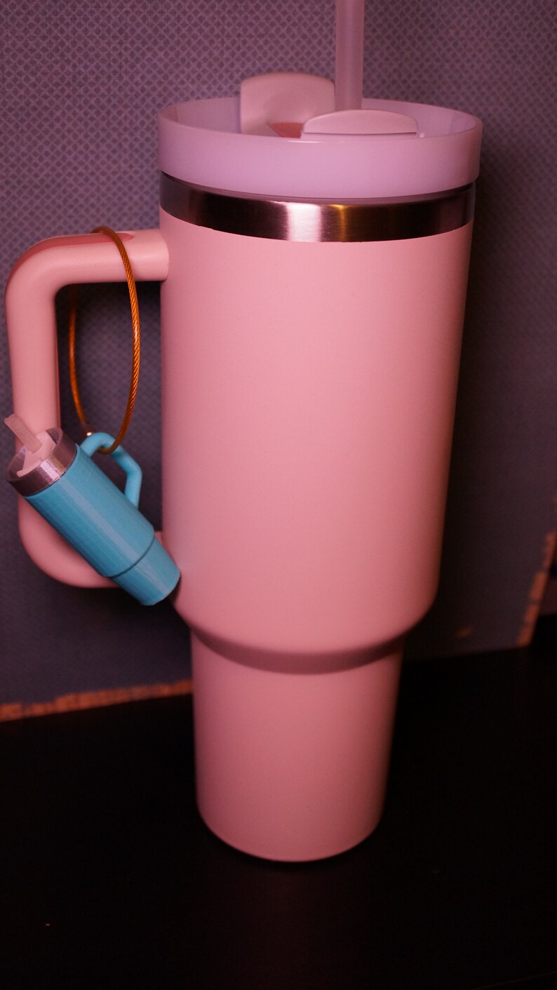 May include: A pink stainless steel travel mug with a white lid and a straw. The mug has a handle and a small blue object attached to it.