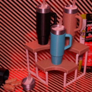 May include: Three insulated travel mugs with lids and handles, in black, brown, and blue, are displayed on a white tiered stand. The stand is on a red and white striped surface. There are three packets of energy drink mix next to the stand.