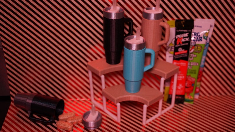 May include: Three insulated travel mugs with lids and handles, in black, brown, and blue, are displayed on a white tiered stand. The stand is on a red and white striped surface. There are three packets of energy drink mix next to the stand.