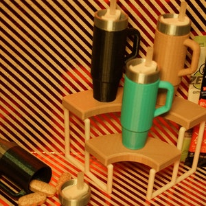 May include: Three stainless steel travel mugs with lids and handles, one black, one brown, and one turquoise, are displayed on a tiered wooden stand. The stand has white metal supports. The mugs are on a striped red and brown background. There are three packets of powdered drink mix on the right side of the image.