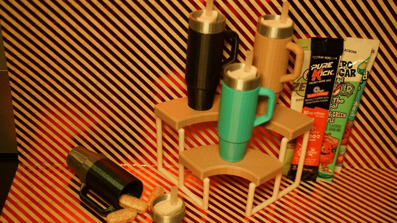 May include: Three stainless steel travel mugs with lids and handles, one black, one brown, and one turquoise, are displayed on a tiered wooden stand. The stand has white metal supports. The mugs are on a striped red and brown background. There are three packets of powdered drink mix on the right side of the image.