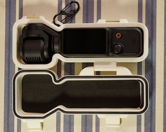 May include: A white plastic case with a black foam interior. The case is open and a black camera gimbal is inside. The gimbal has a small screen and a few buttons.
