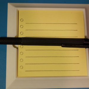 Può includere: Una matita meccanica nera con un accento rosso all'estremità è appoggiata su un blocco note giallo con linee e cerchi. Il blocco note è in una cornice bianca.