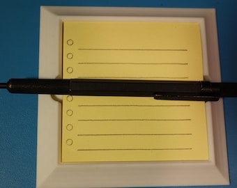 May include: A black mechanical pencil with a red accent on the end is lying on top of a yellow notepad with lines and circles. The notepad is in a white frame.