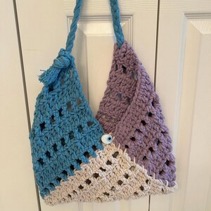 May include: A handmade crochet tote bag in blue, lavender, and cream colors. The bag features a blue rope handle and a decorative button with an eye design. The bag is made of thick yarn with an open weave.