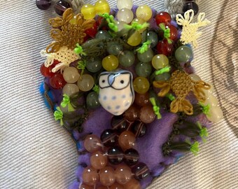 May include: A purple fabric necklace featuring a mix of beads in green, yellow, red and brown. A small ceramic owl with blue and white details is positioned at the centre of the necklace.