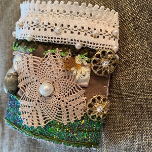 Puede incluir: Un brazalete de tela marrón con purpurina verde, encaje y adornos de perlas. El brazalete tiene dos botones de oro en forma de flor con pedrería.