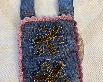 May include: A small denim bag with a purple crocheted border and straps. The bag features two embroidered flower designs with gold and brown beads. The denim has a subtle sparkle, adding a touch of elegance to the handmade accessory.