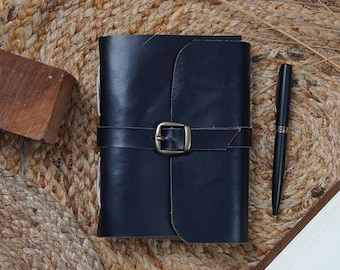 May include: A navy blue leather journal with a buckle closure and a matching pen. The journal is closed with a strap and buckle. The pen is black with silver accents. The journal and pen are on a woven surface.