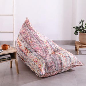 May include: A large beanbag chair with a colourful, patterned design. The fabric features pink, orange, and grey hues with a textured appearance. The beanbag is triangular and rests on a neutral floor. The chair is suitable for indoor use.