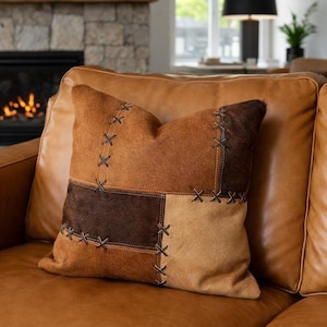 Könnte beinhalten: Ein dekoratives Zierkissen mit Patchwork-Design. Das Kissen weist verschiedene Braun-, Beige- und Dunkelbrauntöne auf, mit sichtbaren Nähten. Es liegt auf einem braunen Ledersofa.