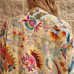 Puede incluir: Kimono beige con estampado floral. El diseño presenta grandes flores coloridas en tonos rojos, azules, amarillos y rosas, sobre un fondo beige. El diseño del kimono es visible desde la espalda.