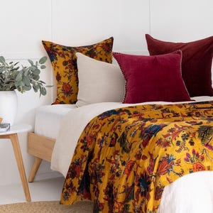 Könnte beinhalten: Ein Bett mit einer senfgelben, floral gemusterten Steppdecke und passenden Kissenbezügen. Zusätzliche Kissen sind in Creme und Burgunderrot. Das Bettgestell ist aus hellem Holz, und ein kleiner Beistelltisch mit einer Pflanze ist sichtbar.