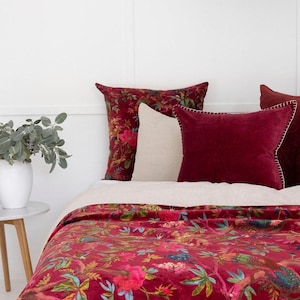 May include: A bed featuring a red floral patterned duvet cover and coordinating cushions. The cushions are in shades of red and cream. A white vase with greenery is on a small side table.