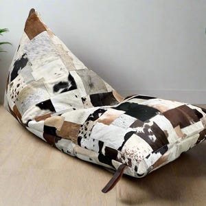 May include: A patchwork beanbag chair in a triangular shape. The chair is made of various cowhide pieces in shades of brown, black, and white. A brown leather strap is attached to the side. The chair is on a light wood floor.