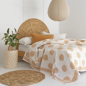 May include: A bedroom scene featuring a bed with a woven headboard and a white and tan patterned quilt. An orange pillow rests on the bed. A round jute rug and a patterned side table are in the foreground. A beige paper lantern hangs from the ceiling.