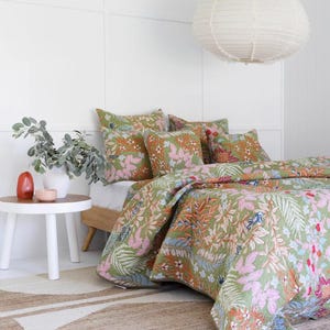 May include: A bed is dressed with a green floral quilt and matching cushions. The quilt features a colourful botanical pattern with pink, orange, and blue accents. A white paper lantern hangs above the bed, and a small side table is nearby.