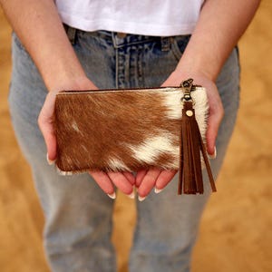May include: A brown and white cowhide clutch bag with a zip and a brown leather tassel. The rectangular wallet is held by a person wearing blue jeans and a white top.
