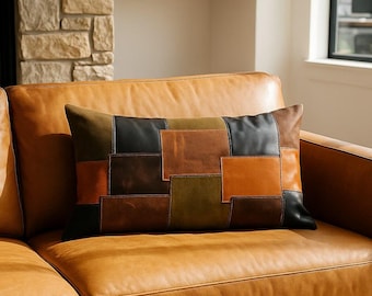 May include: A rectangular decorative pillow with a patchwork design in shades of brown, black, and olive green rests on a tan leather sofa. The pillow's design features rectangular leather pieces sewn together, creating a textured surface.