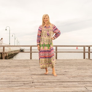 Könnte beinhalten: Ein langes Kleid mit Blumenmuster und bunten Streifen. Das Kleid hat lange Ärmel und eine Quaste am Ausschnitt. Das Kleid wird barfuß auf einem Holzsteg getragen.