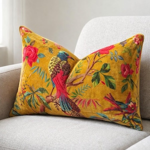 May include: Mustard yellow cushion with a colourful bird and floral pattern. The design features a bird on a branch with red roses and green leaves. The cushion is placed on a light grey sofa.