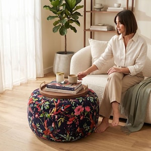 May include: A round, upholstered ottoman featuring a dark navy blue fabric with a vibrant, multi-coloured botanical and bird print. A circular wooden tray with gold-toned metal handles sits on top, holding two books and a small, light-coloured candle.