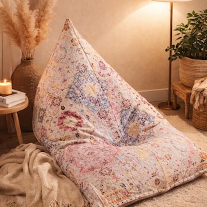 May include: A large, teardrop-shaped beanbag chair with a colorful, floral pattern in shades of pink, blue, and yellow. The beanbag sits on a cream-colored rug, creating a cozy and inviting atmosphere. A soft, beige throw blanket is draped nearby.