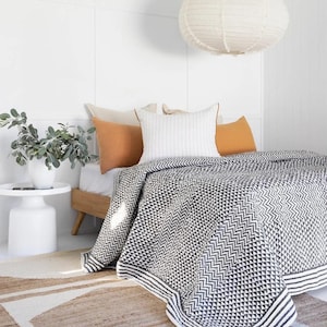 May include: A bed featuring a black and white geometric patterned quilt, orange and white pillows, and a white side table with a vase of greenery. A large white paper lantern hangs from the ceiling.