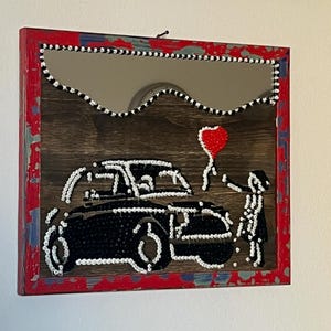 May include: A framed mirror with a red, distressed border. The artwork depicts a black car and a figure holding a red heart-shaped balloon. The mirror's edge is decorated with black and white beads, adding a unique touch to the piece.