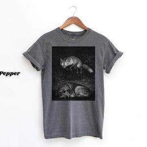 May include: Dark grey t-shirt featuring a black and white illustration of a fox and a wolf. The fox is depicted standing, while the wolf is lying down. The word "Pepper" is printed on the left side of the shirt.