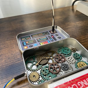 May include: A small metal box with a futuristic control panel design on the lid. The box is open, revealing a collection of gears and clock parts inside.