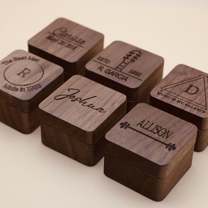 May include: Six wooden ring boxes with personalized engravings. The boxes are made of dark wood and have a square shape. The engravings include names, dates, and symbols like a guitar and a triangle.