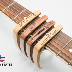 Puede incluir: Cuatro pinzas de capo de madera para guitarra con nombres personalizados y una cita "Siente las notas, vive la música". Las pinzas de capo están en un mástil de guitarra.