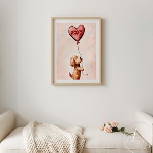 May include: A framed print with a watercolour illustration of a dog holding a heart-shaped balloon. The red balloon has the text "XOXO" on it. The artwork has a soft, pink background and is in a light wood frame.