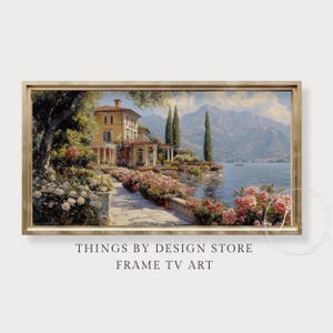 May include: A framed landscape painting depicts a villa with a red-tiled roof, surrounded by lush gardens and a lake. The artwork features a pathway lined with pink and white flowers, with mountains in the background. The frame is a light gold color. The text at the bottom reads "THINGS BY DESIGN STORE FRAME TV ART."