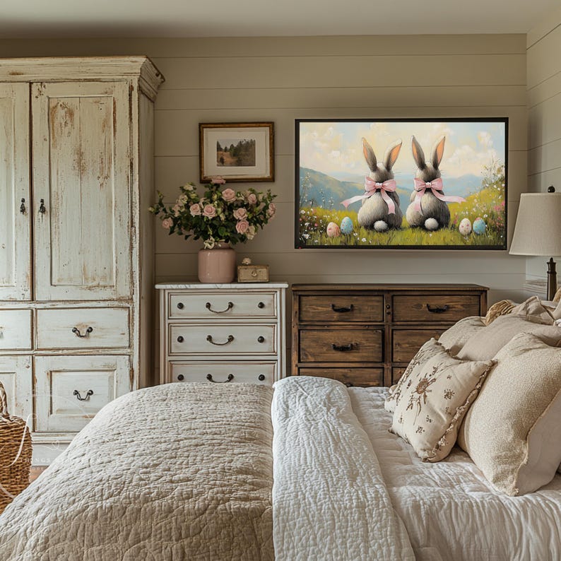 K&ouml;nnte beinhalten: Ein Schlafzimmer mit einer wei&szlig;en Kommode, einer Kommode aus Holz und einem Bett mit einer wei&szlig;en und beigefarbenen Bettdecke. Ein gerahmtes Bild von zwei Hasen mit rosa Schleifen, die auf einem grasbewachsenen H&uuml;gel mit Ostereiern sitzen, h&auml;ngt an der Wand &uuml;ber den Kommoden.