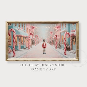 May include: A framed digital art print depicts a snowy street scene with candy canes, buildings, and a nutcracker soldier. The buildings are painted in pastel colors, with pink bows and wreaths. The artwork is labeled "THINGS BY DESIGN STORE FRAME TV ART."