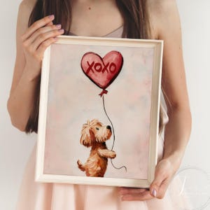 May include: A framed print showing a brown dog holding a red heart-shaped balloon with "XOXO" on it. The artwork has a soft, painterly style with a light pink background. The image is in a white frame.