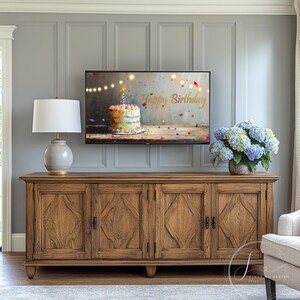 May include: A wooden entertainment console with four doors and a gray wall behind it. A television is mounted on the wall, displaying a birthday cake with a lit candle and the words "Happy Birthday". A lamp with a white shade and a vase of blue hydrangeas are on the console.