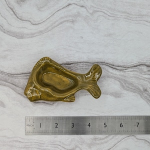 May include: A ceramic fish-shaped dish in a mottled olive green color. The dish is approximately 4 inches long and has a small, shallow bowl for holding items. The tail and fins are detailed, and the dish has a glossy finish.