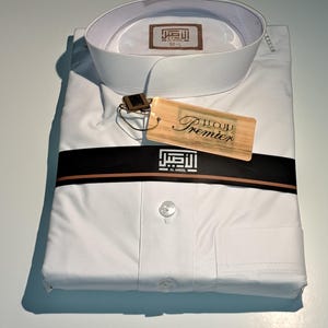 May include: White long-sleeve shirt with a high collar and a single button closure. The shirt is folded and has a brown and black label with the text "Premier" and "Al-Ageel" in Arabic.
