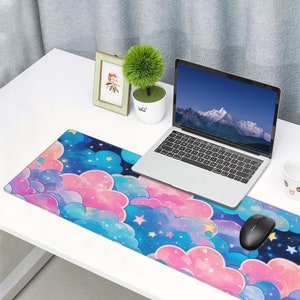 Puede incluir: Un ratón de ordenador negro sobre una alfombrilla de ratón de temática de nubes azules y rosas con estrellas, sobre un escritorio blanco con un portátil, una pequeña planta en maceta y un marco de fotos.