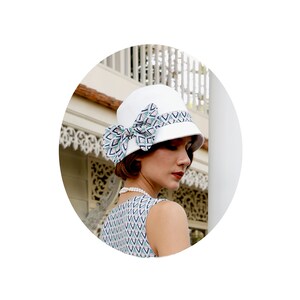 May include: A white cloche hat with a geometric patterned bow. The hat is tilted slightly to the side and the bow is positioned on the top of the hat.